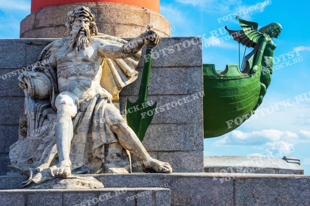 Фотография демонстрирует скульптурные элементы Ростральных колонн на Васильевском острове в Санкт-Петербурге: статую Нептуна и корабельный ростр на фоне ясного голубого неба. Эти скульптуры символизируют морскую мощь и историческое значение Петербурга как важного морского порта. Изображение может использоваться в учебных материалах, культурно-исторических изданиях и как элемент дизайна, отражающий архитектурное наследие города.